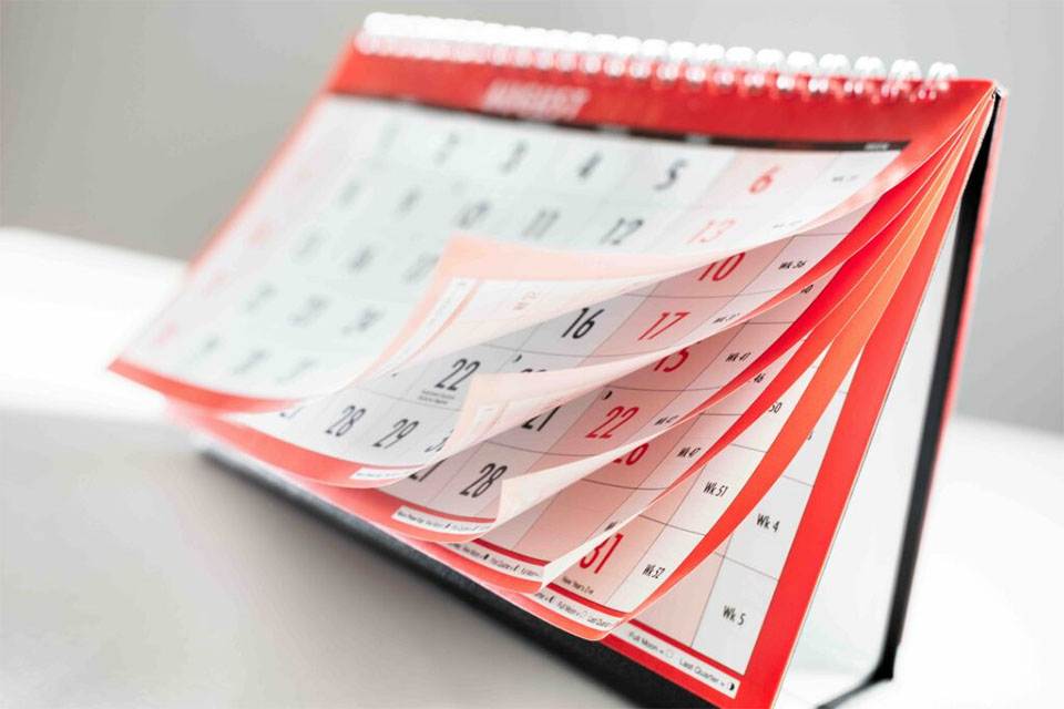 A close-up photo of a desk calendar with several pages flipped, revealing multiple months. The calendar has a red border and sits on a white surface.