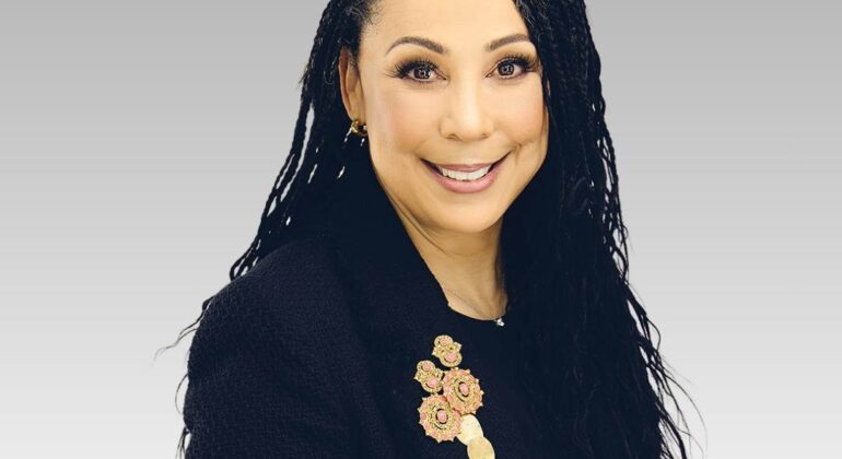 A woman with long, dark, wavy hair wearing a black blazer adorned with a gold and pink floral brooch smiles at the camera against a light gray gradient background, capturing the warmth of sunny afternoons.