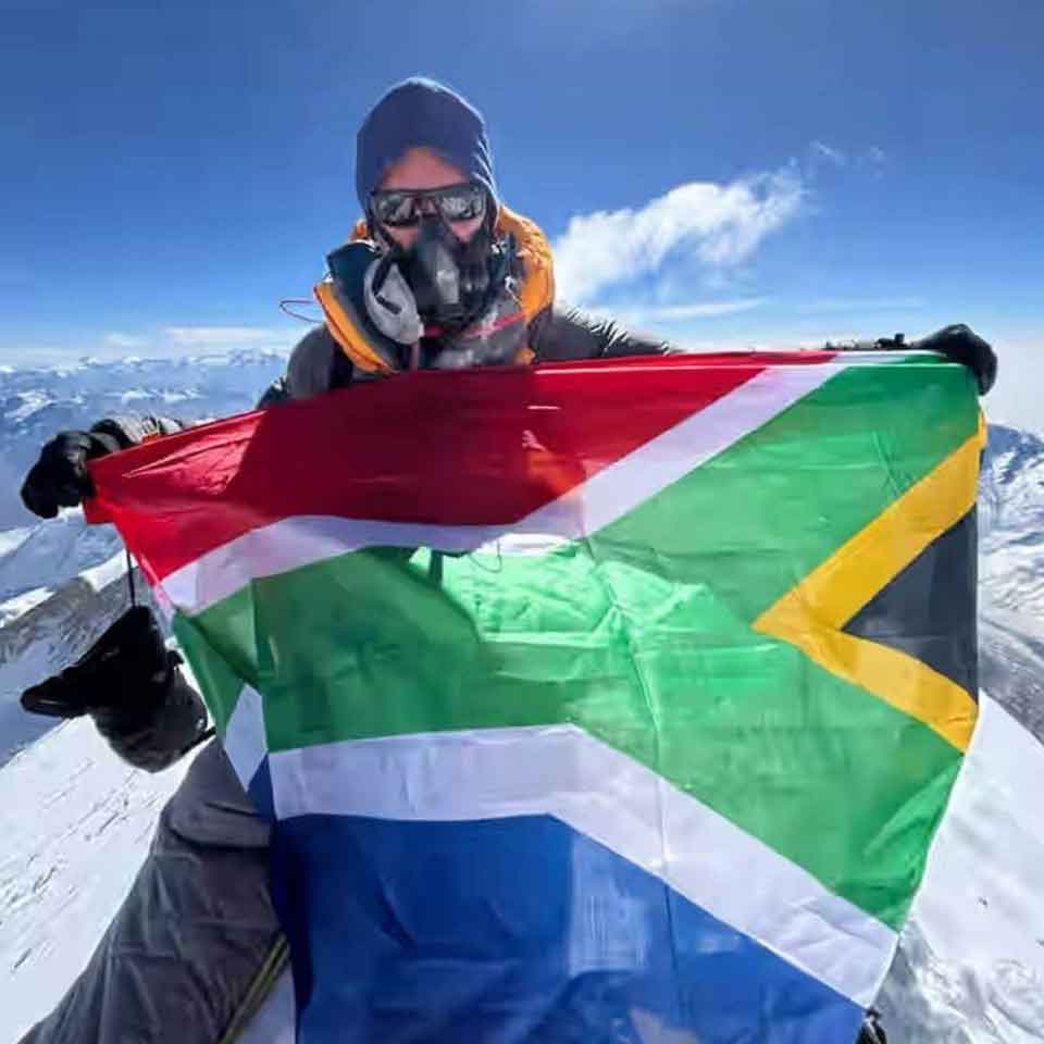 A person wearing cold-weather gear and an oxygen mask holds the South African flag atop a snowy mountain peak, with a clear blue sky and distant mountains in the background.
