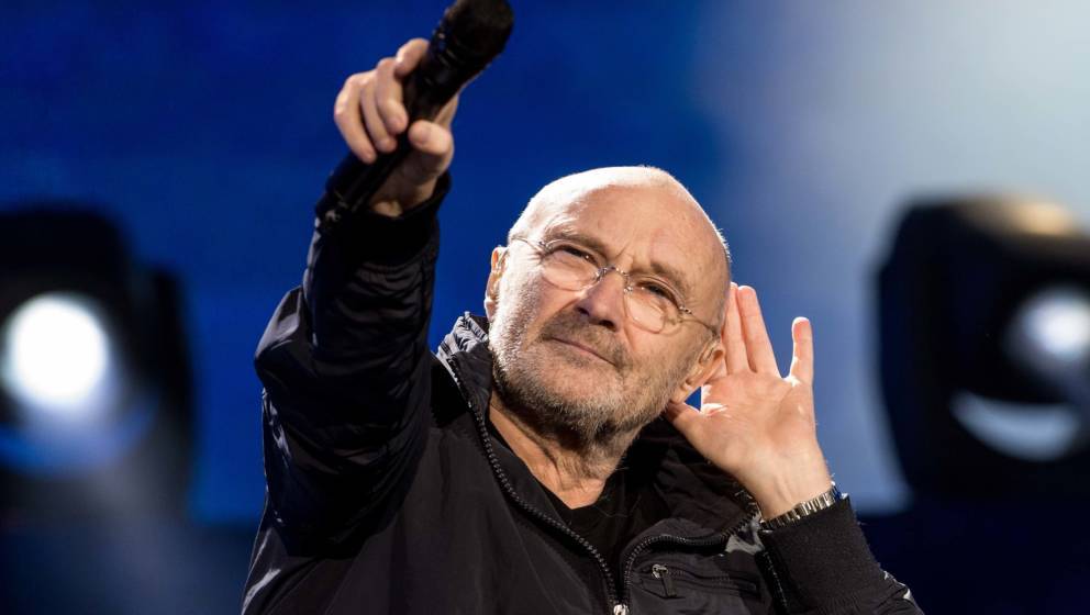 A man with glasses and a beard, dressed in a dark jacket, holds a microphone up while gesturing to his ear on stage during the Matric event, with bright stage lights and a blue background behind him.