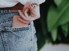 A person wearing blue denim jeans is placing a brown wallet into their back pocket. Their hand is partially visible, and they are wearing a white top and a bracelet. Green leaves are blurred in the background.