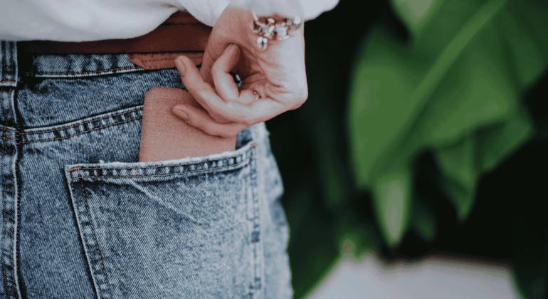 A person wearing blue denim jeans is placing a brown wallet into their back pocket. Their hand is partially visible, and they are wearing a white top and a bracelet. Green leaves are blurred in the background.