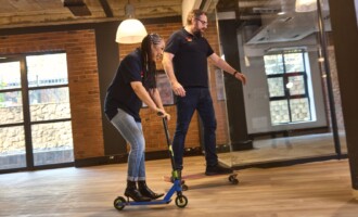 Two people ride in an office; one rides a blue scooter and the other rides a skateboard. Both wear black shirts and jeans, smiling as they move on a wooden floor with brick walls and large windows in the background.