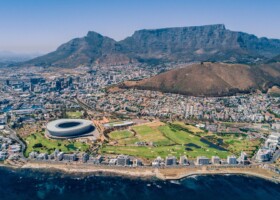 Cape Town. Where city, ocean and mountains meet. Aerial view of Cape Town, South Africa, featuring the coastline, Cape Town Stadium, and the city with Table Mountain and Lions Head in the background under a clear blue sky.